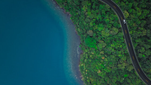 Top view aerial shot of a white car driving on a coastal road between lush green forest and turquoise ocean water, concept of scenic nature landscape representing travel and exploration.	