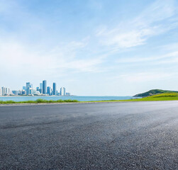 Fototapeta premium An empty asphalt road extends toward a coastal cityscape, with blue sea, green hill and clear sky in the view.