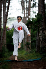 Young girl practicing karate jump in white gi with red belt during outdoor training on a forest path, focused and determined © Nikola Spasenoski