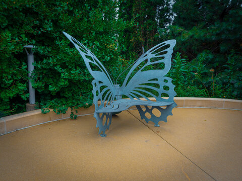 Unique metal butterfly wing bench sculpture positioned outdoors in a lush green garden plaza with stone paving