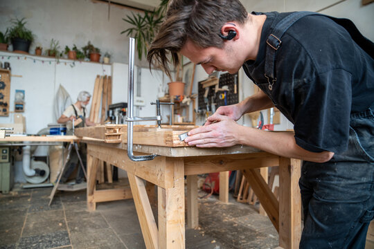 Confident handyman in restoration workshop at table working on old wooden window frame renovation, man blue-collar worker professionally processing wooden frame corners with chisel instrument.