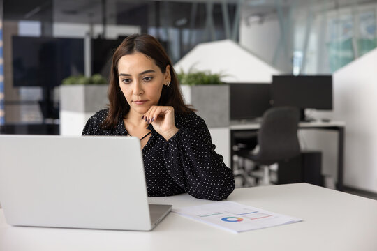 Focused Middle-Eastern businesswoman sits at desk in modern office, analyzing data on laptop, reviewing analytics or financial report, planning strategy, prepare online presentation slides for meeting
