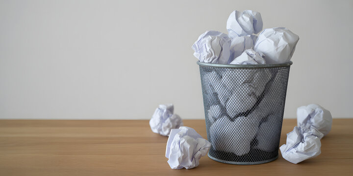 Metal mesh trash bin filled with crumpled white paper balls on wooden desk interior view.