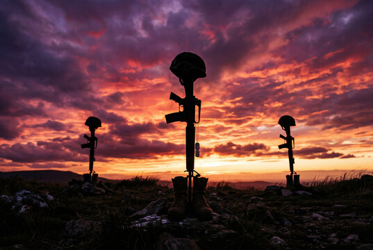 Three military rifles with helmets and boots standing on a dark grassy field at a colorful sunset