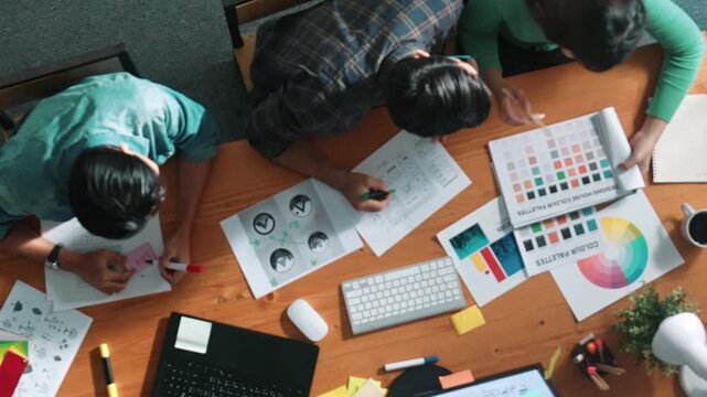Top view of manager writing idea while team standing and giving comment. Professional designer design logo while looking at color palette and writing idea down in paper with color marker. Symposium.