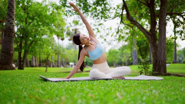 Happy young Asian woman practicing yoga and stretching her shoulders in a park, sitting on an exercise mat on green grass