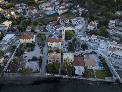 Aerial view of the Church of Saint Spyridon surrounded by residential buildings, lush trees, and the coastline, Argassi, Greece.