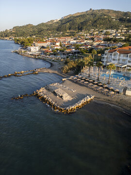 Aerial view of the historic bridge ruins on the beach in Argassi, featuring coastal resort buildings, clear sea, and lush hills in the background in Argassi, Greece.