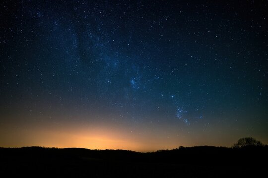 Rural landscape silhouette under brilliant milky way starry sky, warm orange horizon glow over forested hills at night
