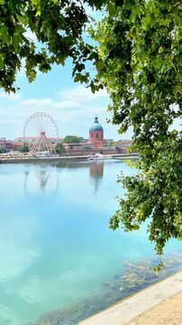 scenic view of toulouse garonne river with ferris wheel and dome framed by green foliage under blue sky