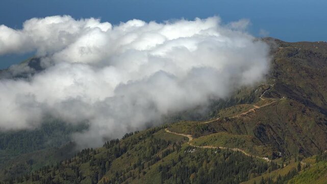 Time lapse clouds moving along a mountain road near the upper border a forest beside rugged slopes.