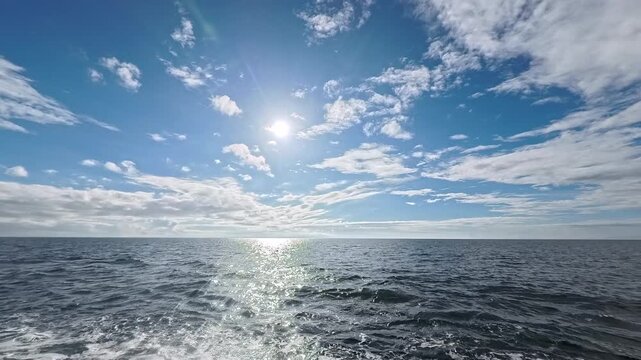 Cinematic real-time wide angle video of brilliant dazzling sun blazing in clear blue sky over ocean with silver reflection