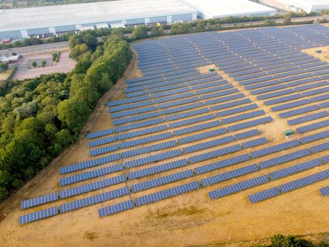 Aerial view of a large solar farm with rows of photovoltaic panels set in a dry grassy field bordered by trees and industrial warehouses in Little Irchester, England, United Kingdom.