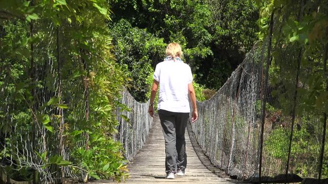 Blonde woman walking across wooden suspension bridge lush forest adventurous travel.