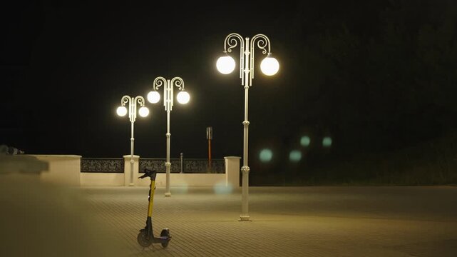 Electric scooter parked on city promenade under street lanterns at night.