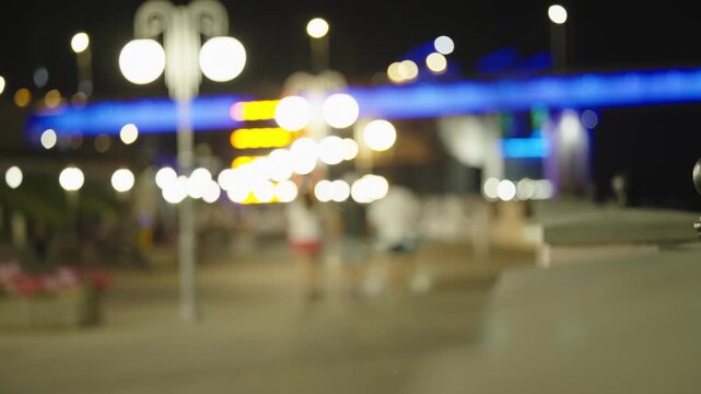Blurry night cityscape with people walking on illuminated city embankment.