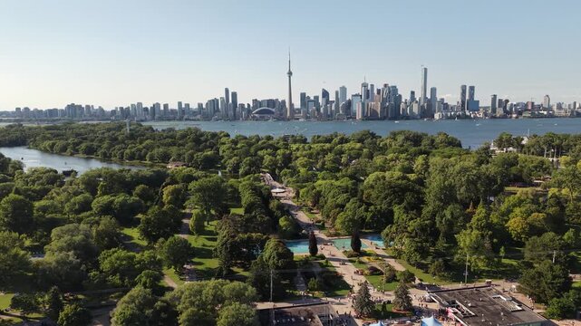 Aerial view tracking right over the lush green gardens, paths, and fountains of Toronto Islands with the iconic downtown city skyline and CN Tower beyond