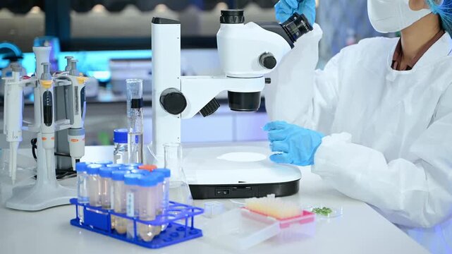 Focused researcher in protective lab gear examines scientific specimens using a stereo microscope in a modern biotechnology laboratory.