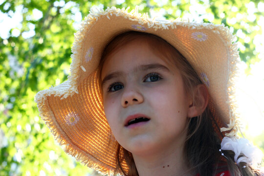 A young girl with light brown hair wearing a straw hat. She has a curious expression and is surrounded by greenery in a bright outdoor setting.