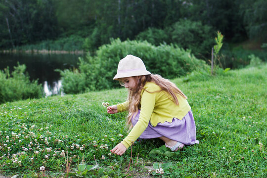 A young girl with long brown hair, wearing a yellow sweater and a pink hat, crouches on green grass, picking flowers near a calm lake surrounded by trees.