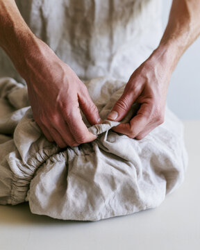 Folding laundry fitted sheet close up view showing hands tucking elastic corner into soft beige linen on clean tabletop with natural texture and copy space