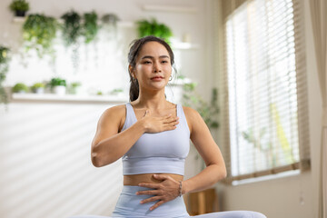 Fototapeta premium Woman yoga meditation breathing home wellness exercise calm indoor plant Asian woman practicing yoga breathing exercise home with calm expression bright room with plant decor and window light