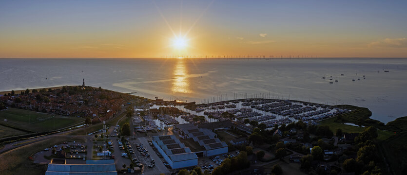Aerial view of the harbor in Hindeloopen at sunset, featuring sailboats in the marina, historic rooftops, and wind turbines on the horizon over the IJsselmeer. Hindeloopen, Friesland, Netherlands.