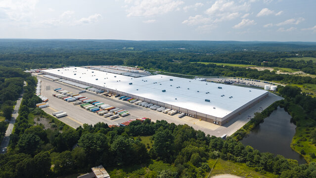 Aerial view of a large distribution center with numerous semi-trucks parked in the loading bays, surrounded by lush green forest and a small pond in Plainfield, Connecticut, United States.
