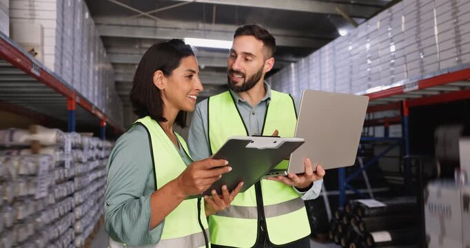 of Hispanic logistic professional colleagues comparing paper and electronic data, using laptop computer and clipboard in warehouse space, talking, discussing inventory