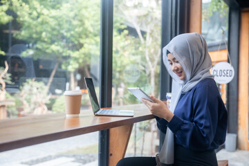 Fototapeta premium Muslim woman using tablet and laptop while working at cafe table with coffee cup