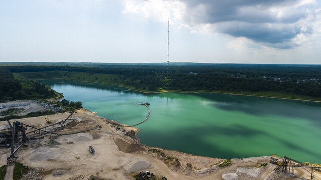 Aerial view of a gravel factory with industrial machinery and a large turquoise quarry lake surrounded by dense forest in Middle Island, New York, United States.