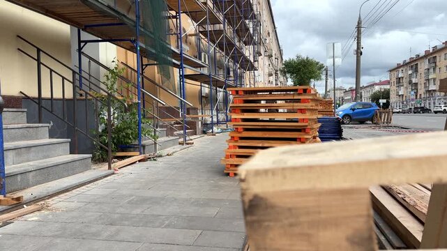 Scaffolded walkway with concrete steps and stacked wooden pallets along a city sidewalk near an old building, material storage, exterior renovation, urban construction, repair zone.