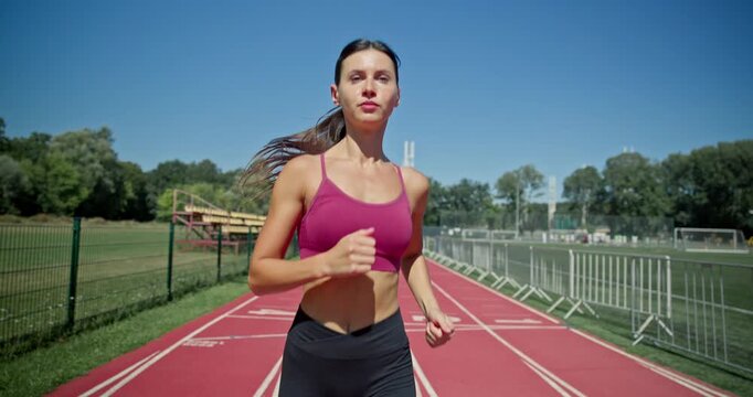 Young athletic girl in sportswear preparing for training at the stadium, warming up and running on the running tracks on a sunny summer day