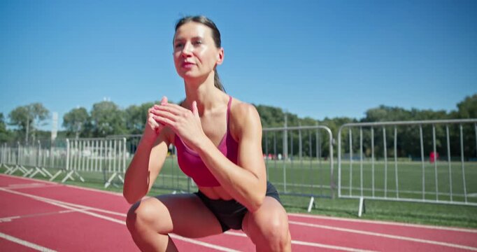 Young athletic girl in sportswear preparing for training at the stadium, warming up and jumping on the running tracks on a sunny summer day