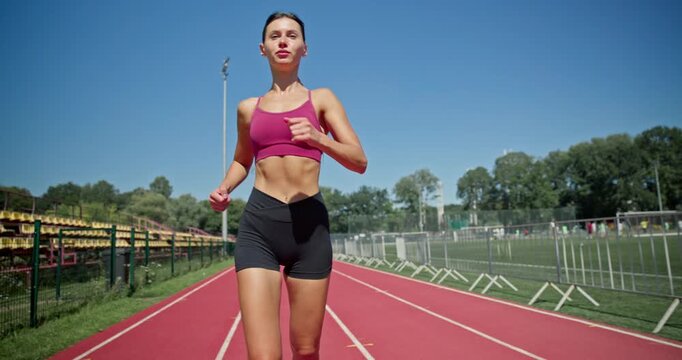 Young athletic girl in sportswear preparing for training at the stadium, warming up and running on the running tracks on a sunny summer day