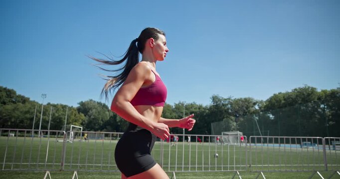 Young athletic girl in sportswear preparing for training at the stadium, warming up and running on the running tracks on a sunny summer day
