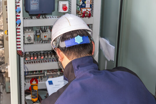Electrician carrying out electrical installation and commissioning work on an open electrical panel in the engine room of a newly built ship, with the worker seen from behind.