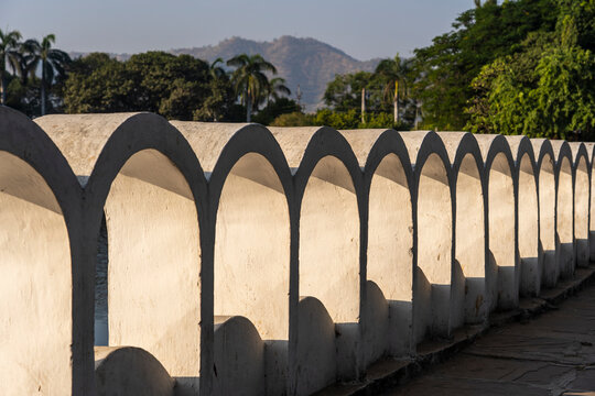 Scenic view of the famous white concrete wall with traditional repeating arches along the walkway at Dudh Talai lake in Udaipur, Rajasthan, India with green trees and hills in the background, closeup