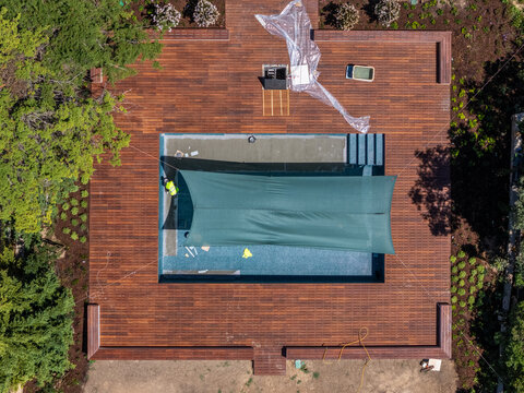 Aerial view of a swimming pool under construction with a green shade cloth covering the water, surrounded by a wooden deck and a worker in a high-visibility vest in Cascais, Lisbon, Portugal.
