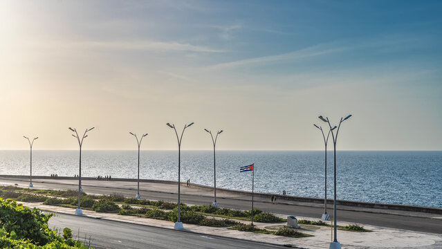 A highway along the ocean shore. People are walking on the Malecon embankment. The flag of Cuba is flying. High street lights, green plants. Blue sky, clouds. Havana