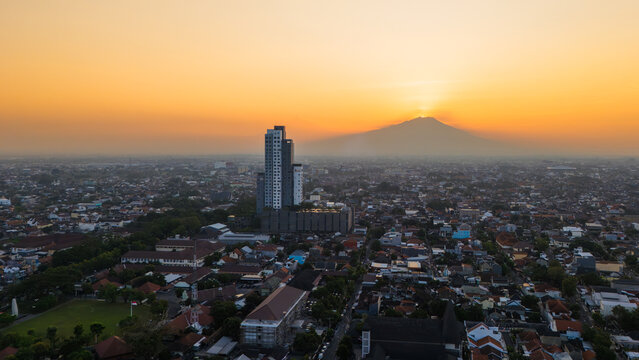 Aerial view of Surakarta city at sunset with Mount Merbabu in the background and a prominent skyscraper rising above the residential rooftops in Surakarta, Central Java, Indonesia.