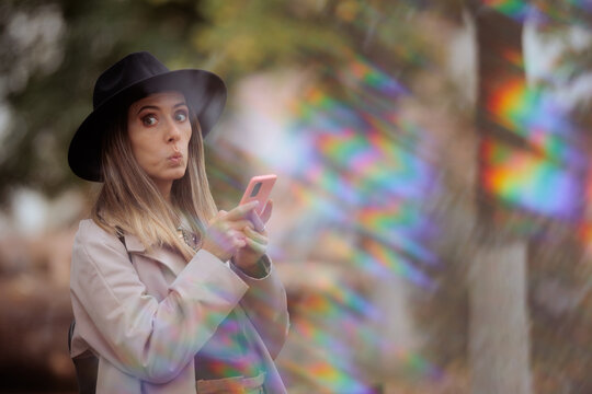 Surprised Young Woman Wearing Fedora Hat Holding Her Pink Smartphone. Shocked girl looks away with wide eyes while checking mobile device