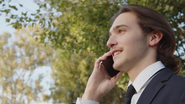 Man in a business suit is walking down the street and talking on the phone. A young man in a suit talking on a smartphone, slow motion. A smiling businessman in the city talking on a cell phone.