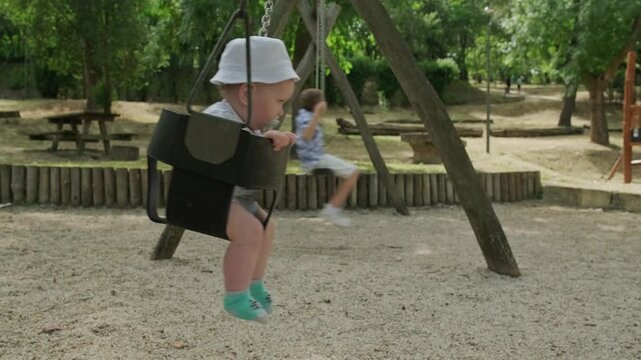 Baby with his older brother swinging in a park on a nice summer day. Two adorable kids on a playground swinging on a swing on a warm spring day. Caucasian children outdoors on the playground.