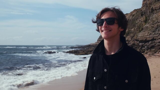 Happy, gorgeous man walks down the beach and glances at the camera. Smiling young brown-haired man with rocks and ocean in the background.  An attractive guy in sunglasses walks through nature.