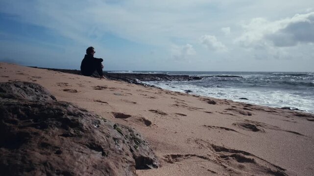Thoughtful handsome young man sits on the empty sandy beach on the summer day.  Guy is sitting on sandy beach on a sunny day.  Man with a romantic mood  on the beach contemplating the beautiful ocean