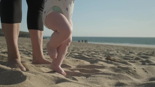 Baby legs in close-up taking the first steps. Toddler takes the first steps with caring mother. Mom teaches her baby to walk on a sandy sea beach. Kid takes first steps with the support of his mother.