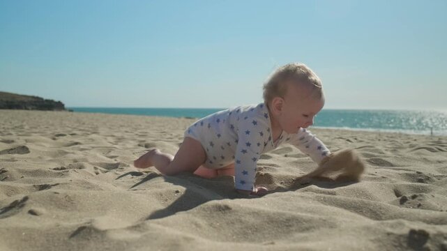 Active, cheerful toddler crawls along a sandy sea beach on sunny summer day. Slow motion: Baby crawling on the sea beach on sunny day. Little boy crawls on sand beach against background of a blue sea
