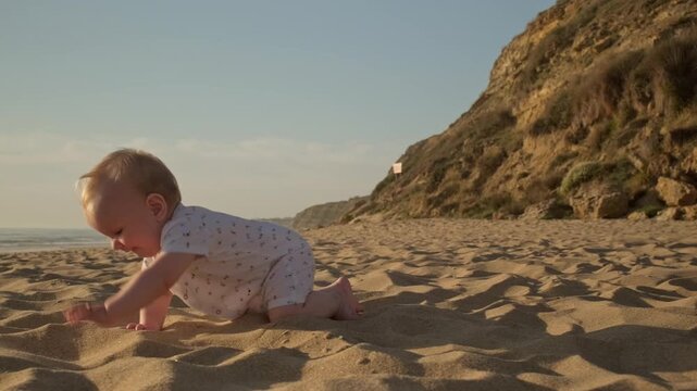 Active, cheerful toddler crawls along a sandy sea beach while sunset. Slow motion: Baby crawling on the sea beach on sunny summer day. Little boy crawls on sand beach against background of a blue sea