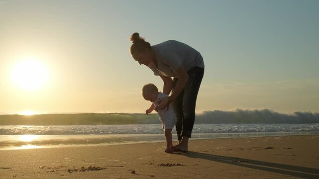 Toddler takes the first steps with caring mother. Mom teaches her baby to walk on a sandy sea beach. Kid takes first steps with the support of his mother on nature. Baby is learning to walk, outdoors.